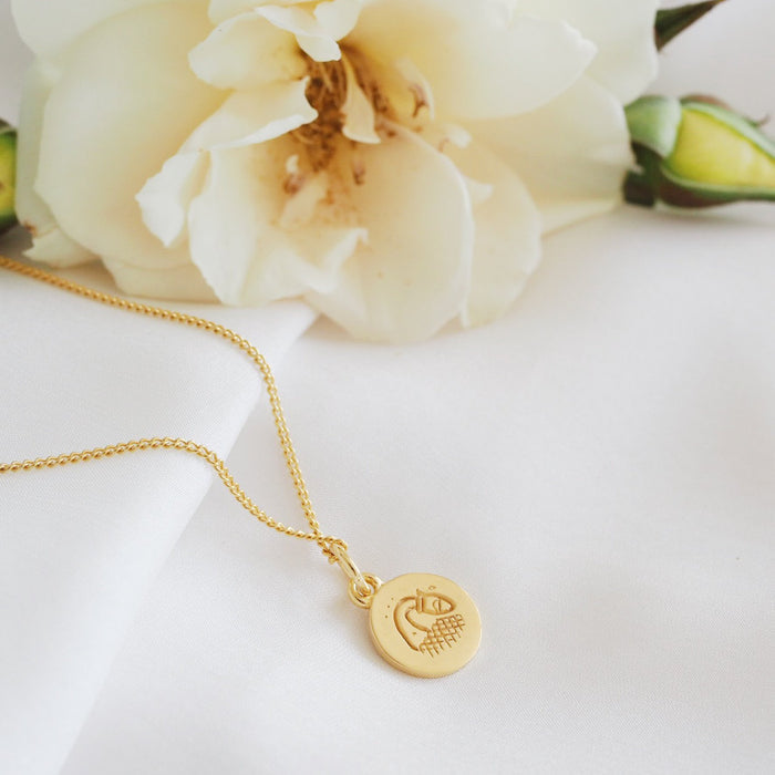 A simple gold necklace featuring a round pendant with a minimalist geometric design, displayed on a white fabric backdrop and a flower.