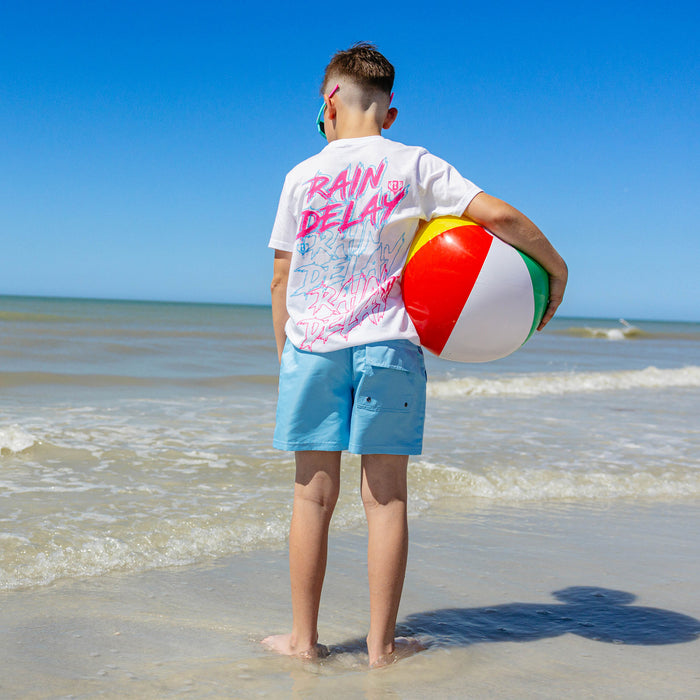 Back view of a young boy in light blue BL101 youth swim trunks and white Rain Delay graphic tee, standing at the water's edge holding a beach ball.