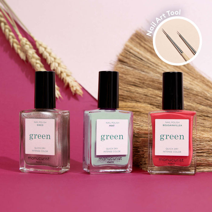 Three bottles of Manucurist Green Flash nail polish, including the Coral Reef shade, are arranged on a pink surface with dried grasses behind them. An inset features a Manucurist Nail Art Brush with dual-sized dotting tips.