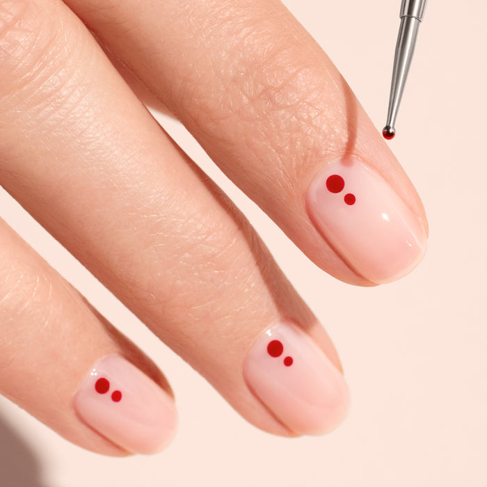 Short, natural nails painted pale pink are adorned with small red dots using the Manucurist Dotting Tool, which features an FSC wood handle for creating minimal dot designs on each nail.