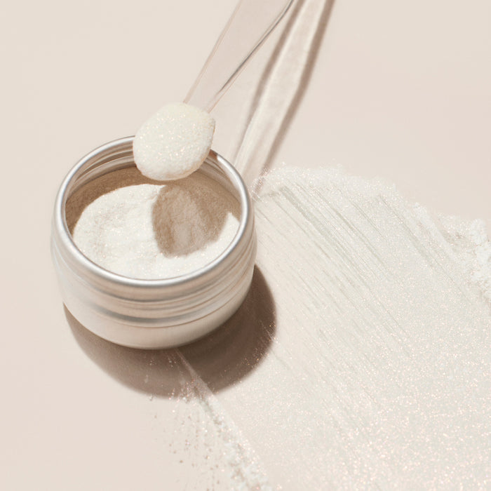 A small metal jar of Manucurist's Glitters Duo Rose Gold Glazed sits on a light surface. A plastic scoop holds some pearlescent powder, with more of the sparkling product spilled and streaked nearby.