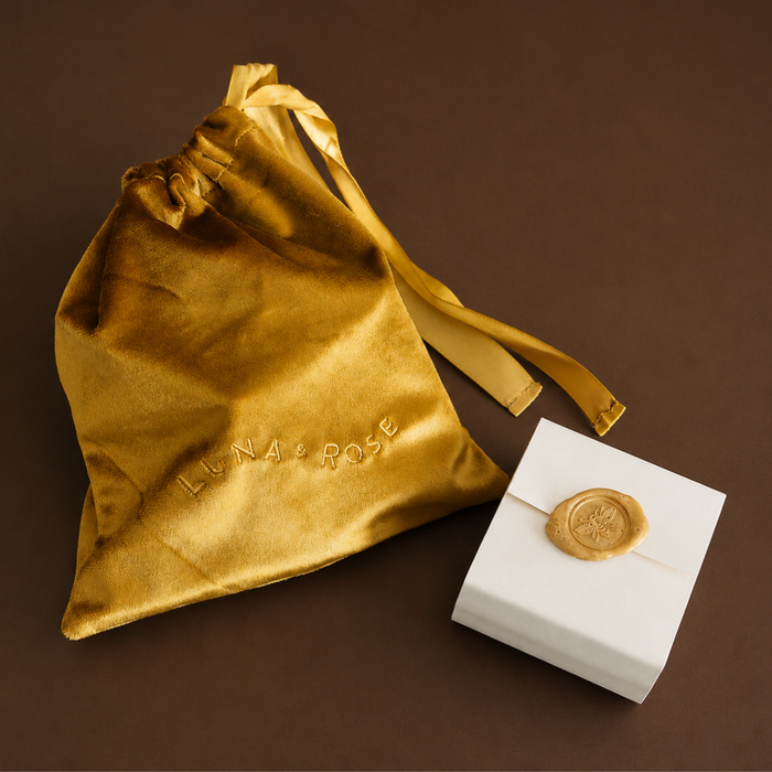 A metallic gold satin drawstring pouch with the brand name debossed on the front, displayed next to a small white gift box sealed with a gold wax stamp on a dark brown background.