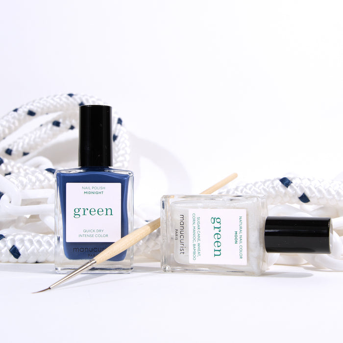 Two bottles of Manucurist Midnight Swim nail polish, one blue and one clear, from the Life Aquatic collection stand on a white surface with a Nail Art Liner Brush. A white rope with blue accents is coiled in the background for a nautical-inspired look.