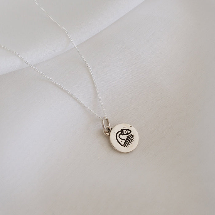 A simple silver necklace featuring a round pendant with a minimalist geometric design, displayed on a white fabric backdrop.