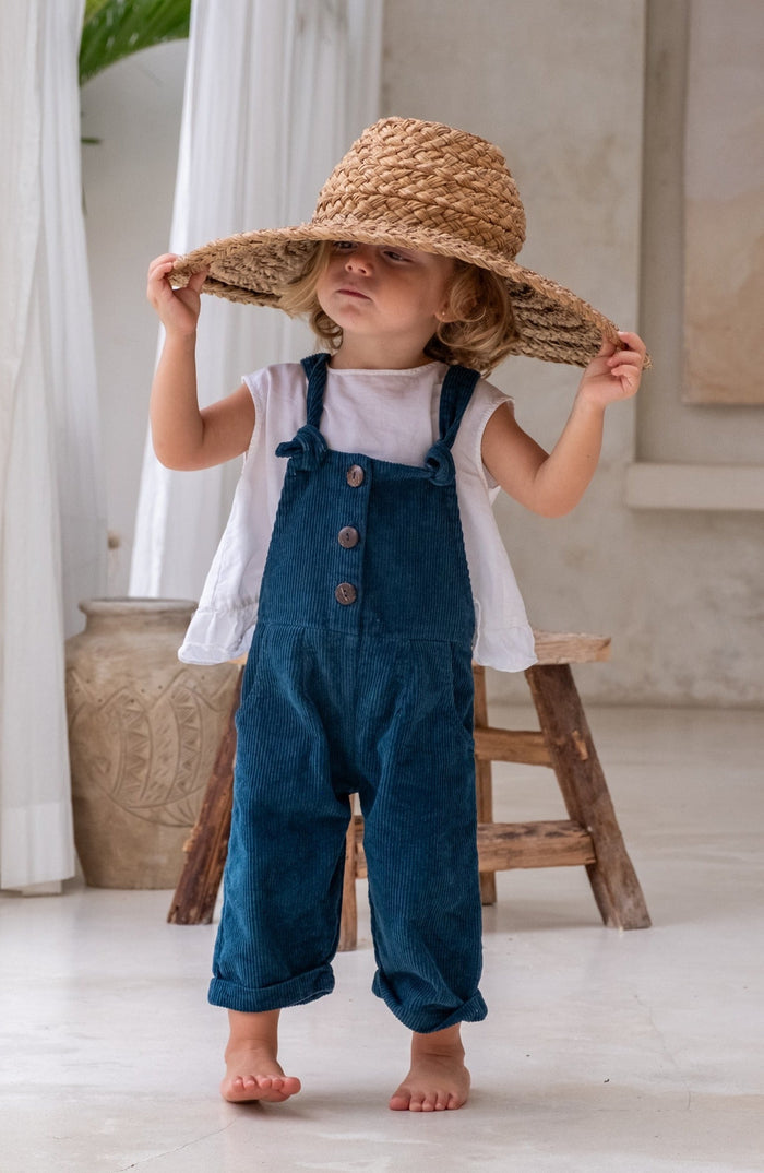 A toddler wearing blue overalls with a white shirt holding her hat