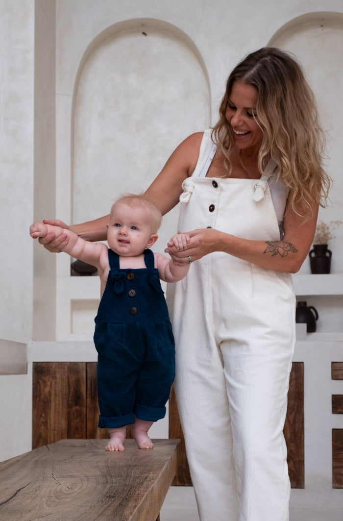 A woman in white overalls helps a baby in blue overalls stand on a wooden bench.
