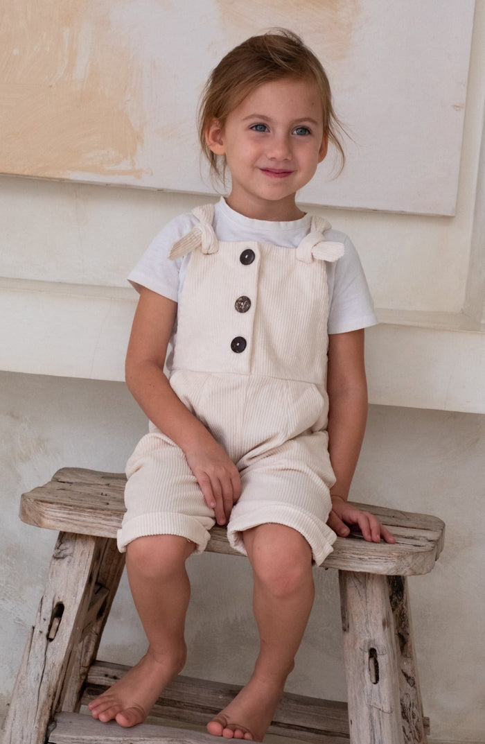 A child in a white overall and white t-shirt sitting on a wooden bench