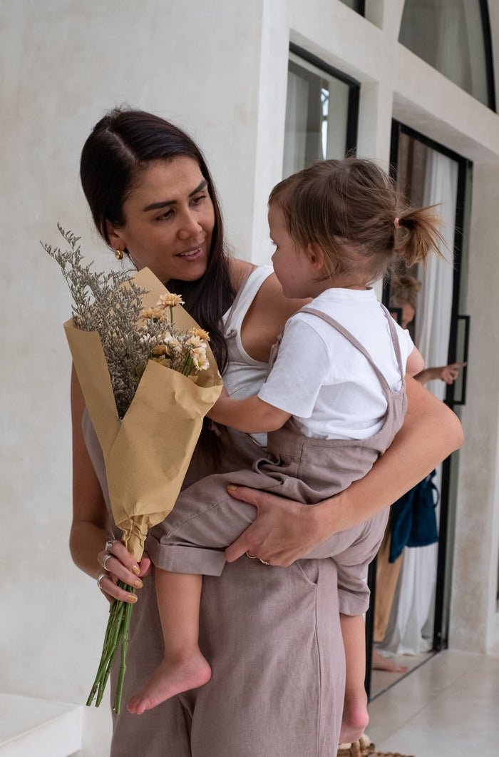 A woman in taupe overalls holds a toddler also in taupe overalls, with the woman carrying a bouquet of dried flowers.