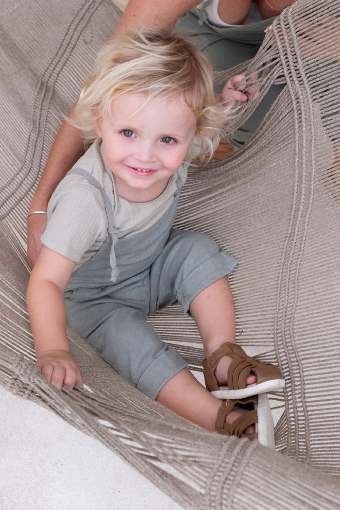 A toddler with blond hair wearing a sage colored overall and brown sandals.