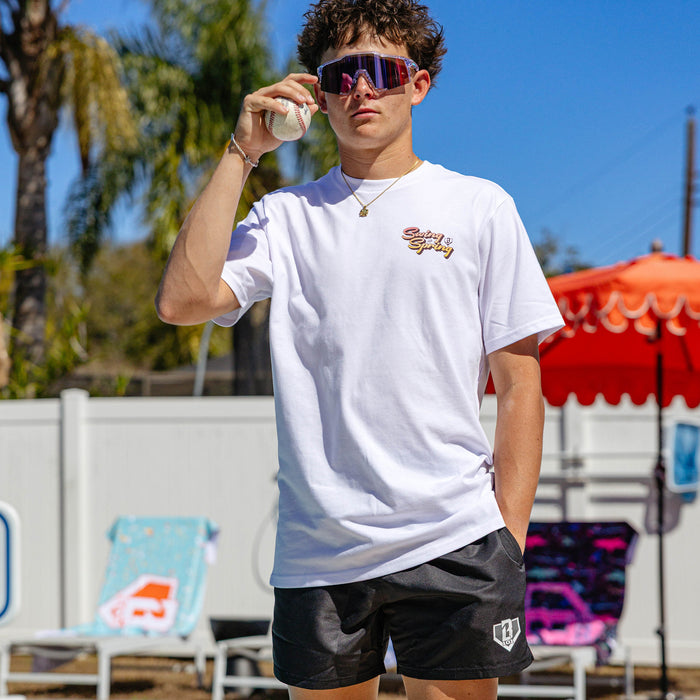 Young man wearing BL101 black swim trunks and a white Swing Into Spring graphic tee poolside, holding a baseball and wearing shield sunglasses.