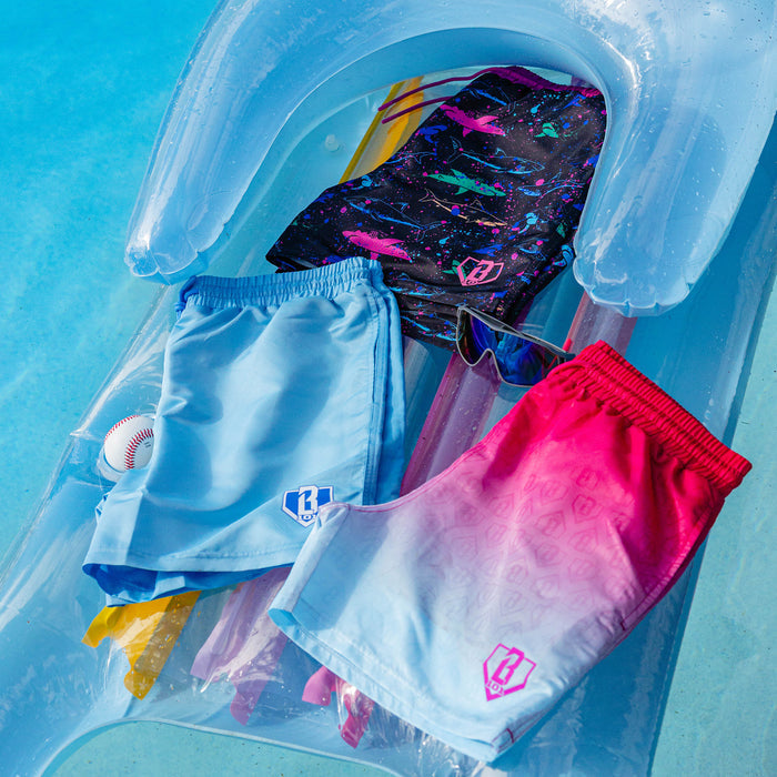 Three BL101 swim trunks in light blue, neon shark, and pink ombre prints piled on a pool float with a baseball and sunglasses.