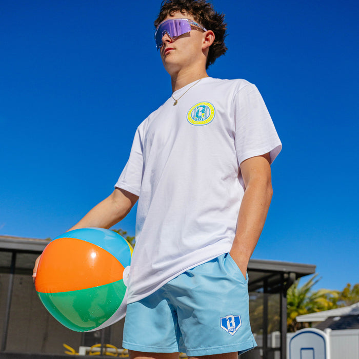 Young man wearing BL101 light blue swim trunks and a white BL101 graphic tee poolside, holding a beach ball and wearing shield sunglasses.