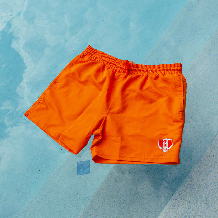Orange BL101 swim trunks with elastic drawstring waistband and white BL101 home plate logo, laid flat on a light blue pool deck.