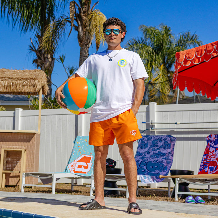 Male model standing poolside in orange BL101 swim trunks, white BL101 tee, dark slides, and blue mirrored sunglasses, holding a colorful beach ball.