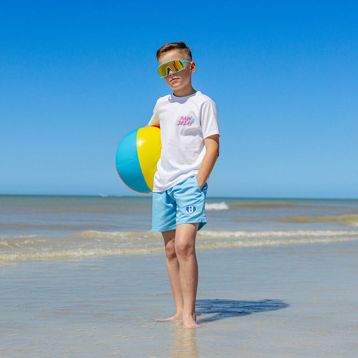 Young boy on the beach wearing light blue BL101 youth swim trunks and a white Rain Delay tee, holding a beach ball while wearing yellow mirrored shield sunglasses.