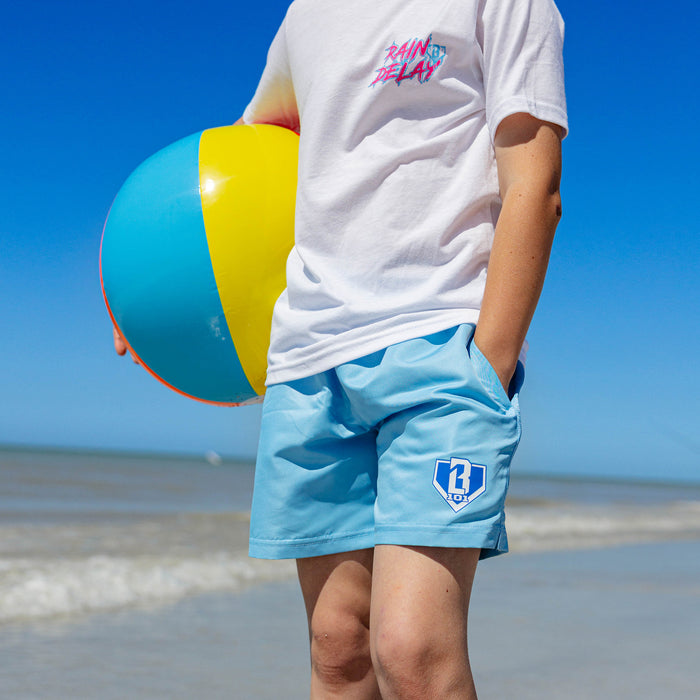 Close-up of light blue BL101 youth swim trunks with blue and white BL101 home plate logo, worn at the beach.