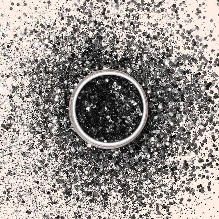 A silver ring rests on a light surface, surrounded by Manucurist Biodegradable Glitter in black and silver, forming a sparkling circular pattern around the ring.