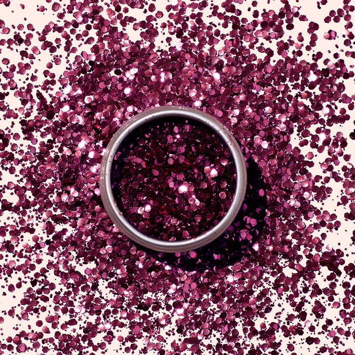 A round metal tin of Manucurist’s Bordeaux Biodegradable Glitter sits on a surface, surrounded by scattered magenta glitter that sparkles in a circular pattern matching the eco-friendly product.