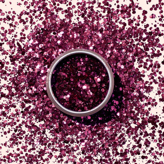 A round metal tin of Manucurist’s Bordeaux Biodegradable Glitter sits on a surface, surrounded by scattered magenta glitter that sparkles in a circular pattern matching the eco-friendly product.