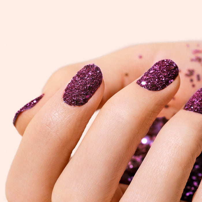 A close-up of a hand with nails painted sparkly purple gel polish. Manucurist’s Bordeaux Biodegradable Glitter is scattered on the skin and soft beige background.