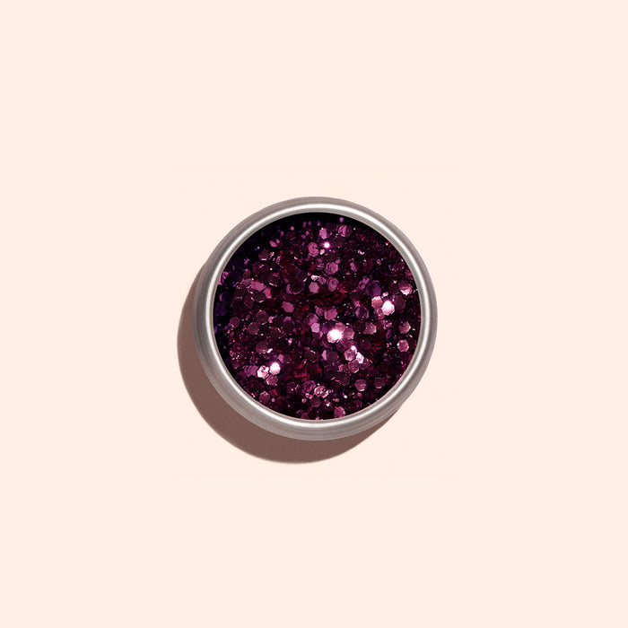 A round, open container of Manucurist Bordeaux Biodegradable Glitter sits centered on a pale pink background, its chunky magenta sparkles catching the light and reflecting shades of pink and purple.