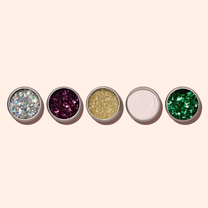 Five small round containers of Manucurist's Bordeaux Biodegradable Glitter are arranged in a row on a beige background, showcasing shimmering eco-friendly glitter eyeshadows in silver, purple, gold, white, and green.