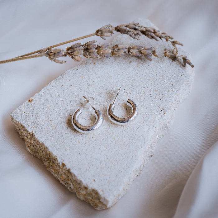 A pair of silver hoop earrings with post backs displayed on a textured beige stone, accompanied by a sprig of dried lavender.