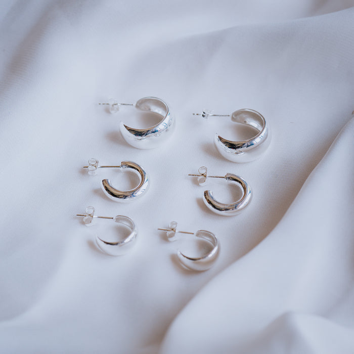 Three pairs of varying-sized silver hoop earrings with post backs arranged on a white fabric surface.