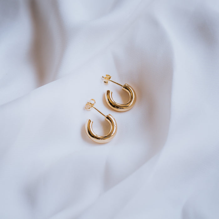A pair of golden hoop earrings with post backs arranged on a white fabric surface.