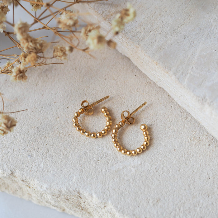 Gold beaded earrings displayed on a textured beige stone with dried flowers.