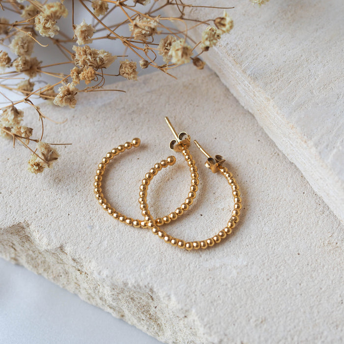 Gold beaded earrings displayed on a textured beige stone with dried flowers.