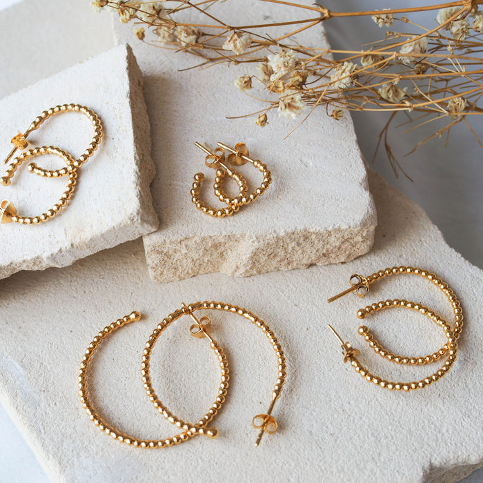 Multiple gold earrings displayed on a textured beige stone with dried flowers.
