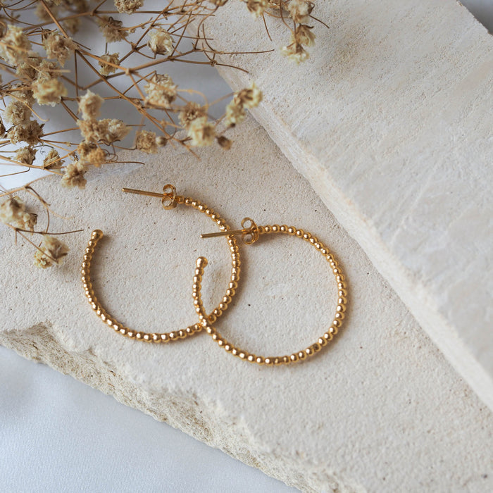 Gold beaded earrings displayed on a textured beige stone with dried flowers.