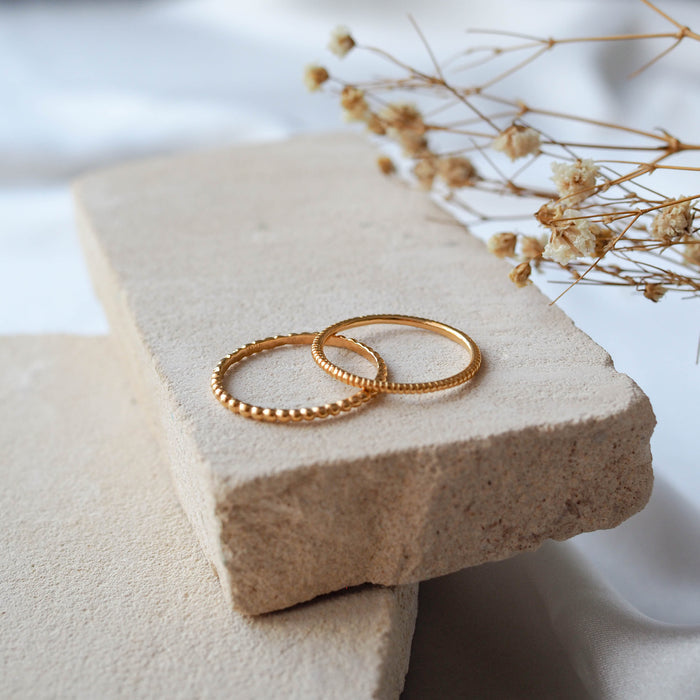 Gold beaded rings displayed on a textured beige stone with dried flowers.
