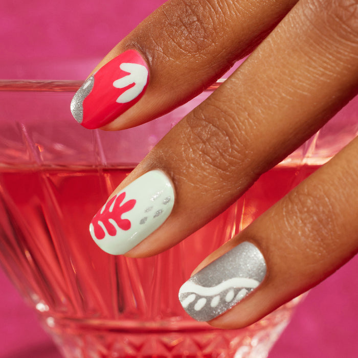 A hand with manicured nails painted in red, white, mint green, and silver using Manucurist’s Coral Reef features abstract wavy designs created with a Nail Art Brush, held above a glass of pink liquid against a pink background.