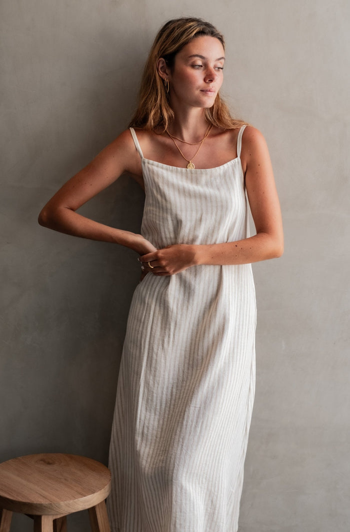 A woman in a striped midi dress, standing in a minimalist space with a wooden stool beside her.
