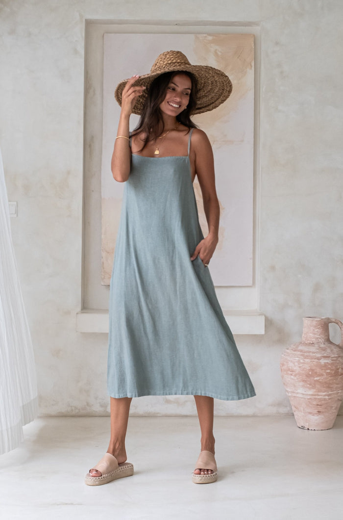 A woman in a light blue sundress and wide-brimmed straw hat, standing in a minimalistic room with neutral-colored decor and pottery in the background.
