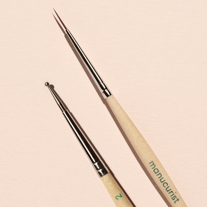 The Manucurist Dotting Tool  Nail Art Liner Brush Duo features light wooden handles and silver tips, and is shown arranged on a pale pink background.