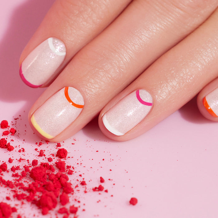 A hand with short, polished nails shows a shimmery light pink base and colored semicircle cuticle accents, created using Manucurist's Dotting Tool  Nail Art Liner Brush Duo. Red powder is scattered on the pink surface nearby.