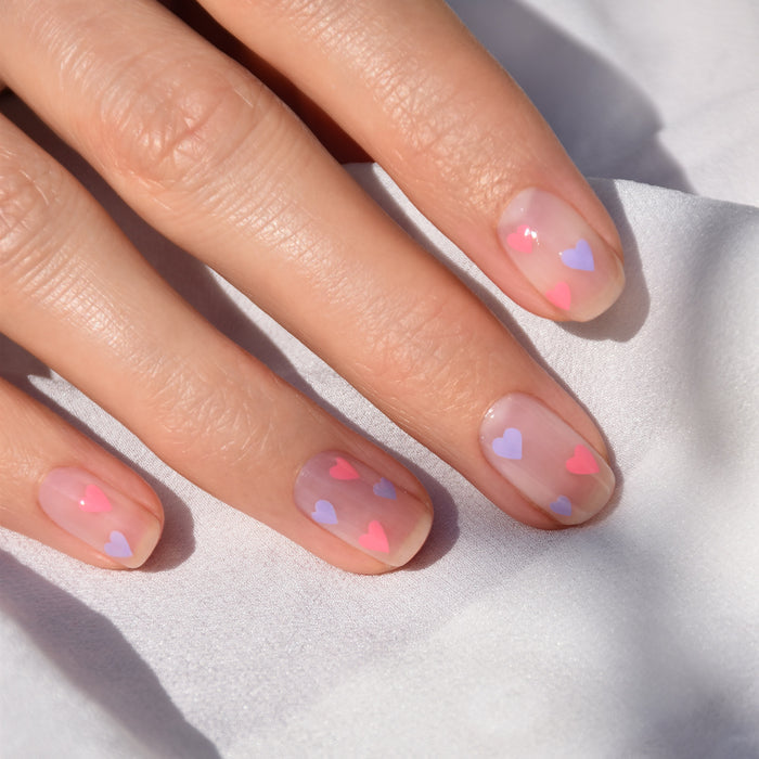 A hand with natural nails features small pink and purple heart designs created using the Manucurist Dotting Tool  Nail Art Liner Brush Duo, shown close-up against a white fabric background.