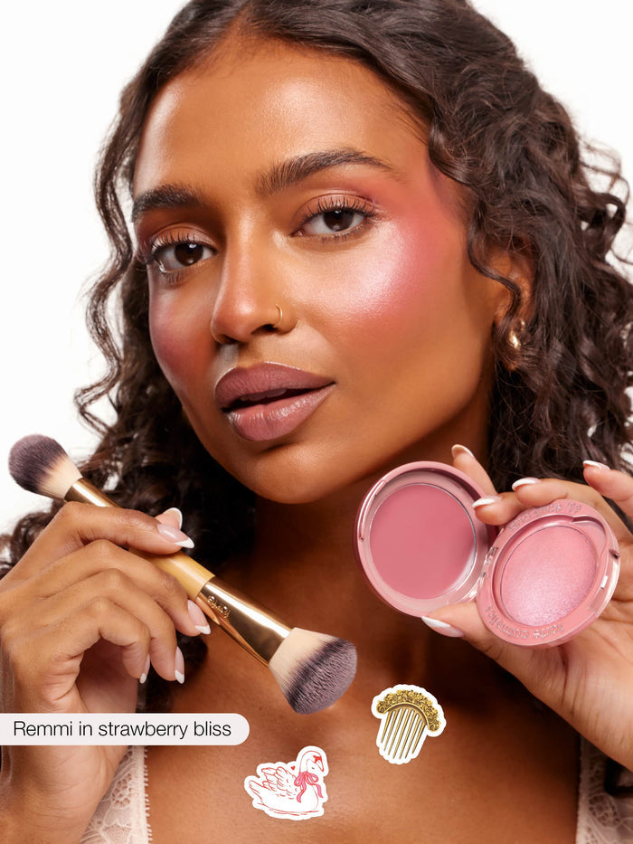 Woman holds an open pink cream blush compact and a gold-handled brush, blending rosy color on her cheeks against a white studio background. Text present: Remmi in strawberry bliss