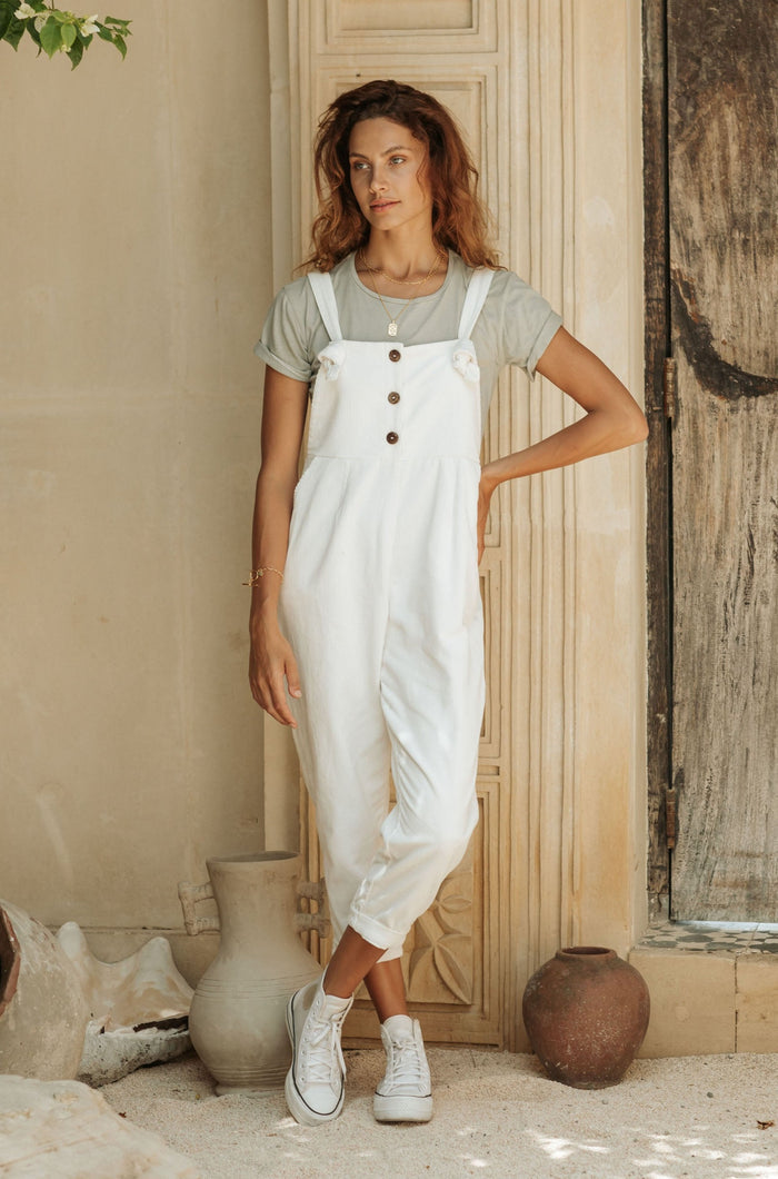 A woman in white overalls and a gray t-shirt, standing against a decorative wooden door with pottery in the background.