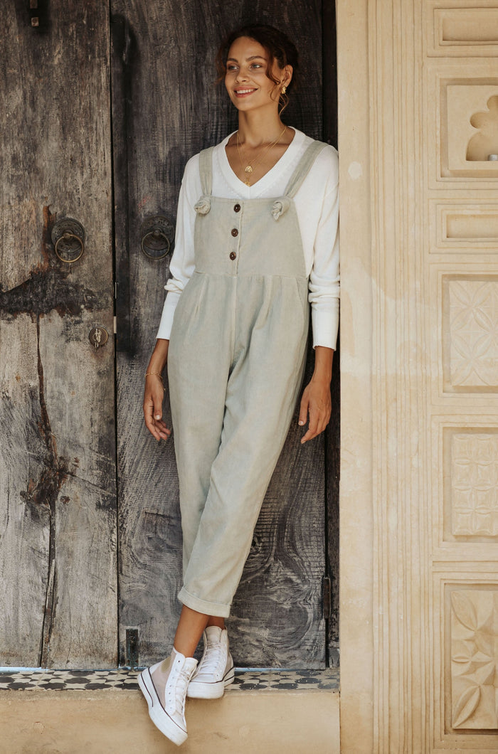 A woman in sage overalls and a white tank top, wearing white sneakers leans against a decorative wall.