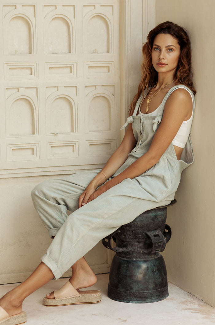 A woman in light green overalls and a white tank top, sitting on a decorative metal stool against a patterned wall.