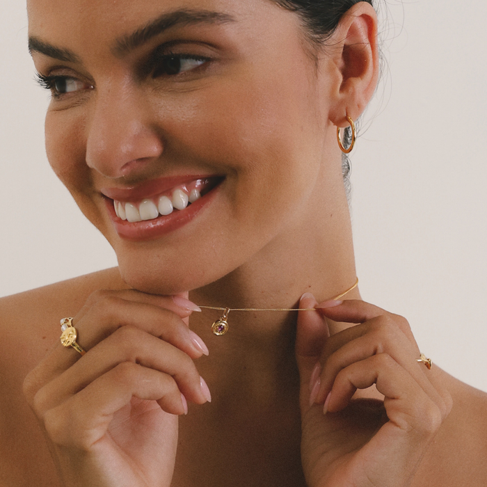 Woman wearing gold jewelry including earrings, necklace, and ring on a neutral background
