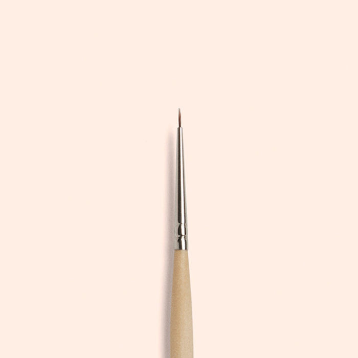 The Manucurist Fine Tip Brush, made with vegan synthetic bristles, a wooden handle, and a silver ferrule, stands vertically against a light beige background—perfect for creating precise nail art.