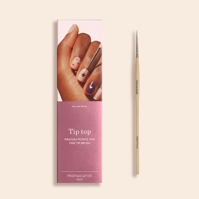 A Fine Tip Brush for nail art with a wooden handle and vegan bristles sits beside its pink box, which shows decorated nails and the text "Fine Tip Brush by Manucurist.