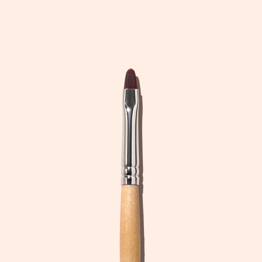 A single Manucurist Flat Brush with a wooden handle and brown bristles is vertically centered on a plain, light beige background, making it ideal for precise manicure and cuticle care.