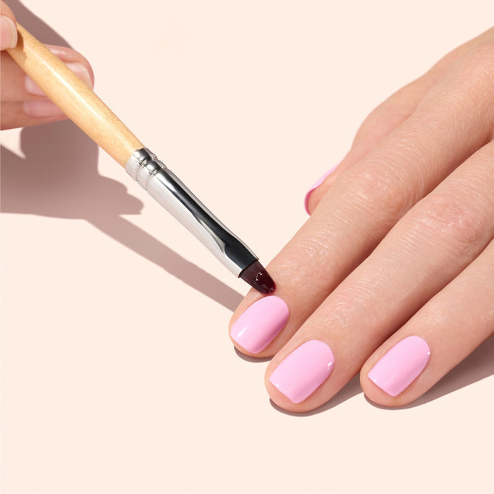 A hand with short, light pink painted nails is being carefully tidied at the cuticles using the Manucurist Flat Brush. The background is a soft beige color.