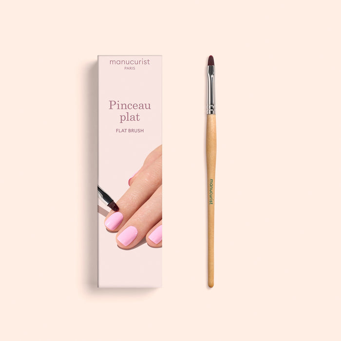 A Manucurist Flat Brush with a wooden handle, ideal for precise manicure and cuticle care, is positioned next to its box, which features an illustration of a hand with pink nails being painted. The packaging displays the brand name "Manucurist" along with "Flat Brush.